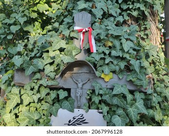 Warsaw, Poland. 31.10.2021 A Jesus Christ On The Cross Metal Figure On The Gray Cross And White And Red Ribbo Among The Poison Ivy With At The Military Cemetery At Powązki During Sunny All Saints Day