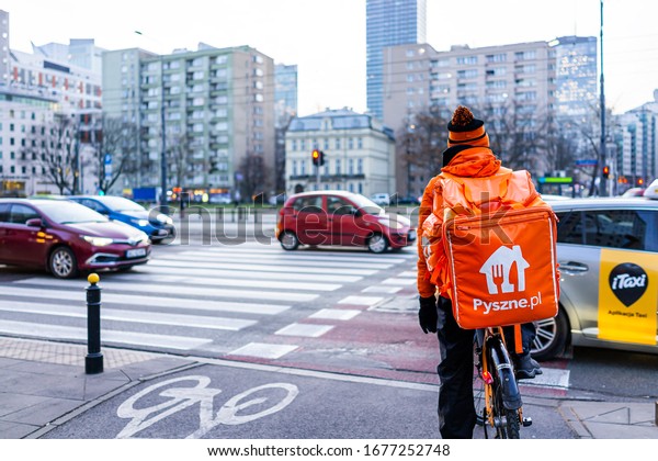 Warsaw, Poland - December 19, 2019: Pyszne.pl\
courier in orange work clothes delivery man delivering online food\
orders on bicycle in\
winter