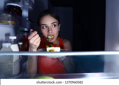 Wary young woman eating food near fridge, view from inside