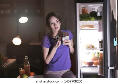 Wary Young Woman Eating Unhealthy Food Near Fridge In Kitchen At Night