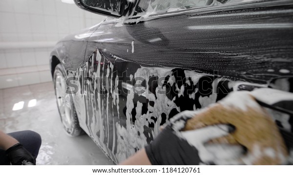 washerman is rubbing surface of black automobile by\
sponge with soap foam in car washing, wearing rubber gloves,\
close-up of hand