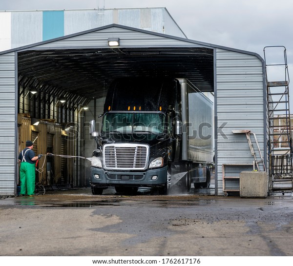 Washers team wash\
a bonnet industrial diesel black big rig semi truck with dry van\
semi trailer at a specially equipped truck wash box for high cab\
trucks and full size\
trailers