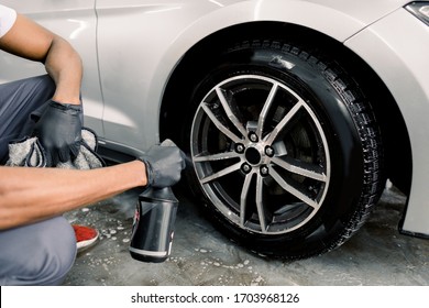 Washing a car by hand, car detailing. Cropped image of hands of young man in protective gloves spraying special cleaning solution on the rim of modern white sport car with at carwash service