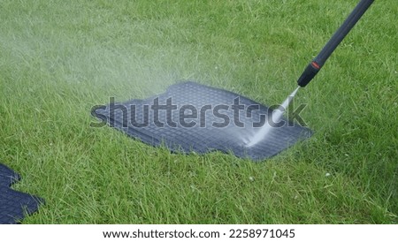 Washing car rubber black mats with manual pressure washer on green lawn. Splashing water scatters and cleans mat on grass