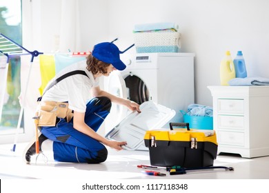 Washing Machine Repair Service. Young Male Technician In Blue Uniform Examining And Repairing Broken Washer Or Tumble Dryer. Maintenance Of Home And Household Appliance. Plumber Apprentice At Work.