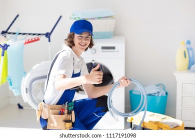 Washing Machine Repair Service. Young Male Technician In Blue Uniform Examining And Repairing Broken Washer Or Tumble Dryer. Maintenance Of Home And Household Appliance. Plumber Apprentice At Work.