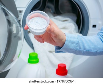 Washing powder detergent, in the background an open washing machine with white laundry. Hand holds a measuring cup of washing powder.