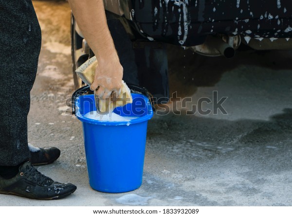 washing sponge\
in hands, blue bucket soap foam. photo of bucket for car wash on\
sunny summer day at home area, Images with car care content. sponge\
in a man\'s hand, close-up\
