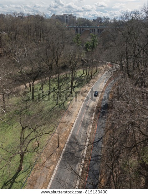 WASHINGTON, DC -\
MARCH 2017: Lone car on lonesome highway in wooded Rock Creek,\
stark trees, distant skyline, road less traveled.  Rock Creek is\
welcome green swath in urban\
setting.