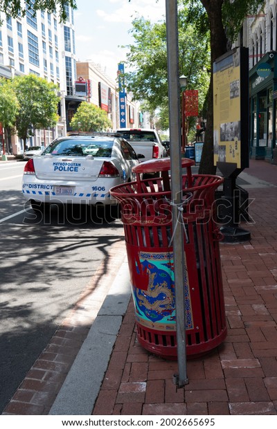 Washington, DC, US - June 23, 2021: Steel trash can\
receptacle secured to sign post for pedestrian foot traffic to\
deposit waste into rubbish bin for weekly collection by city\
government, police car\
