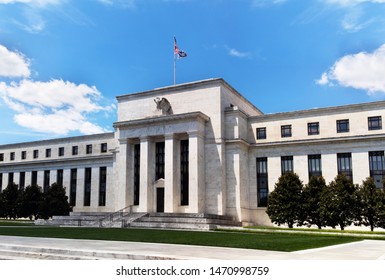 Washington DC, USA - June 22, 2019: Exterior View Of Federal Reserve Board Of Governors Building Facade Entrance With Eagle Stone Statue And American Flags Over A Blue Cloudscape Sky.