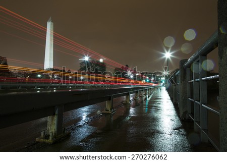 Washington Monument Lit Up at Night in the Rain with Light Trails from Cars. The photo was taken from the Tidal Basin Bridge, showing the reflection of the lights on the sidewalk and pavement.