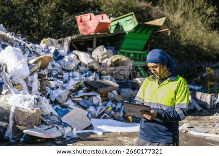 Waste management worker using clipboard to inspect and monitor garbage and trash at a landfill site, ensuring proper waste disposal and recycling procedures
