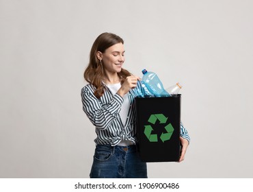 Waste Recycling Concept. Smiling Young Woman Holding Black Container With Plastic Bottles And Green Recycle Sign, Millennial Eco-Friendly Lady Standing Over Gray Background In Studio, Copy Space