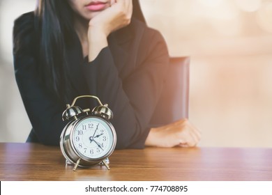 wasting time concept with Asian business woman feeling tired and bored waiting for someone coming late at work and looking at alarm clock on desk  
