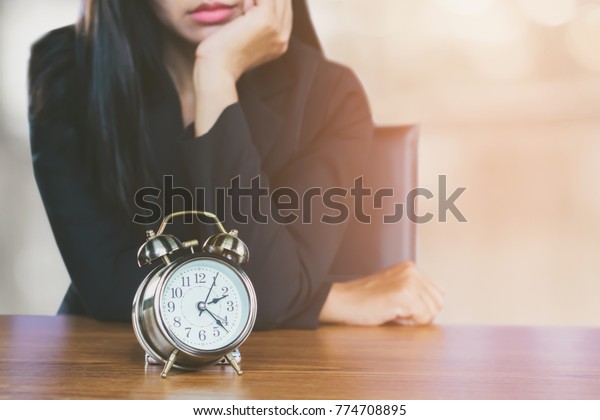 wasting time concept with Asian business\
woman feeling tired and bored waiting for someone coming late at\
work and looking at alarm clock on desk \
\
