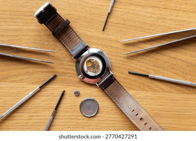 Watch featuring a leather strap and opened back rests on a wooden surface, surrounded by various watch repair tools including tweezers and a battery