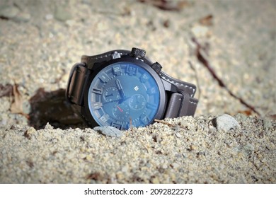 A watch with a leather strap is lying on the sand.