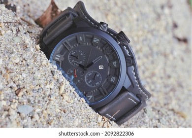 A watch with a leather strap is lying on the sand.