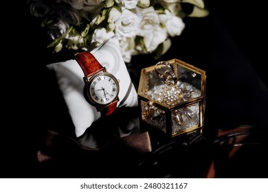 A watch and a ring are displayed on a table. The watch is red and white, and the ring is gold. The table is set with a white cloth and a bouquet of white flowers