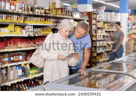 Watchful aged man and woman customers selecting frozen food from market fridge in superstore