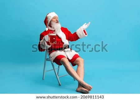Watching football match online. Senior man wearing Santa Claus costume, holding glass of lager foamy beer isolated on blue background . Concept of holiday, event, festival, activity. Copy space for ad