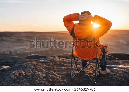 Watching the sunset from a mountain peak while relaxing in a camping chair in an orange jacket as day turns to night