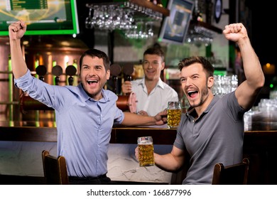Watching TV in bar. Two happy young men drinking beer and gesturing while sitting in bar