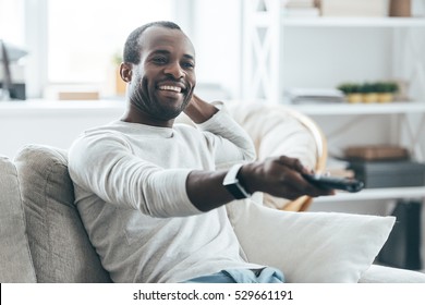 Watching TV At Home. Handsome Young African Man Watching TV And Smiling While Sitting On The Sofa At Home 