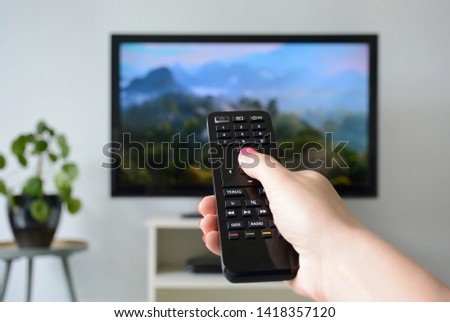 Watching TV. A woman's hand holding the TV remote control with a television in the background. Nature, documentary, tv screen, binge watching