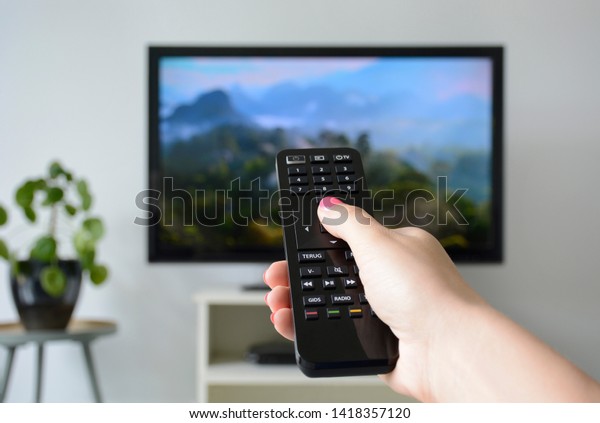 Watching TV. A woman\'s hand holding the TV\
remote control with a television in the background. Nature,\
documentary, tv screen, binge\
watching