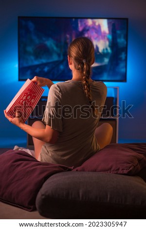 Watching your favorite movies and programs on TV. A young blonde woman sits in front of the TV with a packet of popcorn. Blue neon light. Relax. Hobby. Rest. Favorite TV shows.