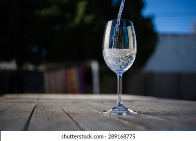 Water Being Poured Into A Wine Glass On A Rustic Table Outside With Natural Light. 