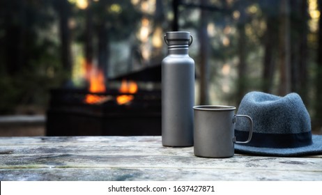 Water Bottle, Hat And Titanium Mug Near The Fire Outdoors. Bushcraft, Adventure, Travel, Tourism And Camping Concept. Copy Space.