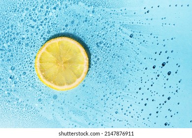 Water with bubbles and round slice of lemon with drops on blue background, flat lay, copy space. Minimal backdrop for cosmetic product or summer travel concept.