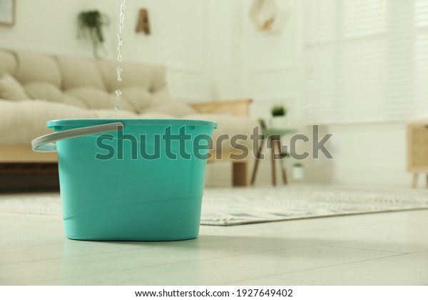 Water dripping from ceiling\
into plastic bucket on floor in living room, space for text.\
Leaking roof