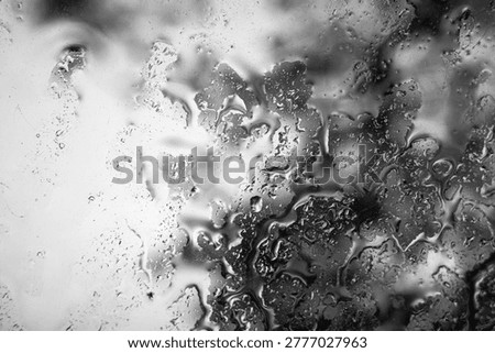 Water droplets gather on a window, distorting the view outside. The scene shows blurred outlines of trees and grass, with clouds overhead during a rainy day.