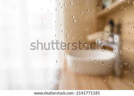 Water drops on wet glass shower door in hotel bathroom with blured bokeh window, sink, faucet and wooden furniture on sunny morning or day. Travel, holiday, vacation, interior design, body care