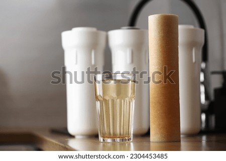 Water filter cartridge used and a glass of rusty water brown coloring on wooden table at kitchen interior. Plastic set with three filter cartridges reverse osmosis water purification system