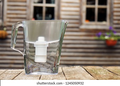 Water Filtration Pitcher Standing On A Rough Wooden Table In Front Of The Windows Of A Country House Outdoors                    