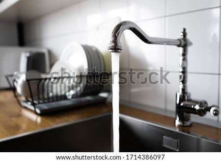 Water flowing out of a kitchen stainless steel tap into the sink. Wasting water by leaving a chrome faucet tap running. Overusing household water. Water misuse in domestic duties and activities.