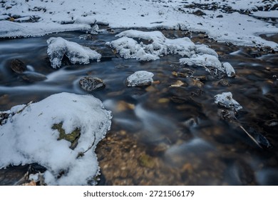 Water flows in a cold stream between rocks and snow. Ice and frozen crystals are visible on the rocks. It is winter in the forest. 
