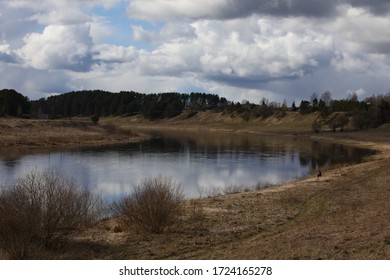 Water Landscape In The Countryside With Wooden Houses On A High Bank.A Bend In A Wide, Calm River With A Fisherman Sitting In The Distance With A Fishing Rod Under A Cloudy Blue Sky In Spring.Russia