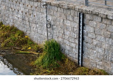 Water level gauge on the Oder beside stone embankment wall with monitoring camera and surrounding riverside vegetation