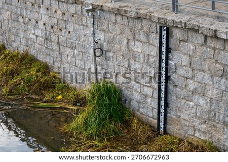 Water level gauge on the Oder beside stone embankment wall with monitoring camera and surrounding riverside vegetation