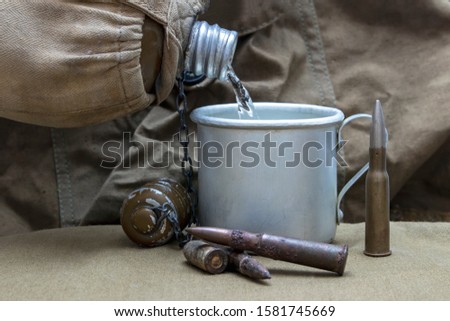 Water is pouring from an old battle flask into a mug. In the background is a cartridge and a combat camping bag.
