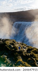 Water pours over dramatic cliffs, creating a stunning Dettifoss waterfall in Iceland. A person stands at the edge, absorbing the serene beauty as mist rises, illuminated by the golden sunlight.