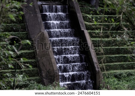 Water from a river creek flows down a wall like stairs, resembling a waterfall. The wall is covered with moss, adding to the natural beauty. The scene is captured on Sandiaoling, Taiwan.