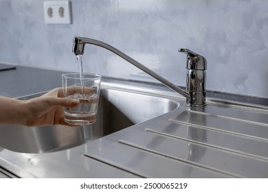 Water is running from the tap in the kitchen. A girl takes water into a glass from the tap in the kitchen