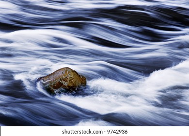 Water Rushing By A Rock In A River Forming A Smooth, Abstract, Painted Appearing Pattern.